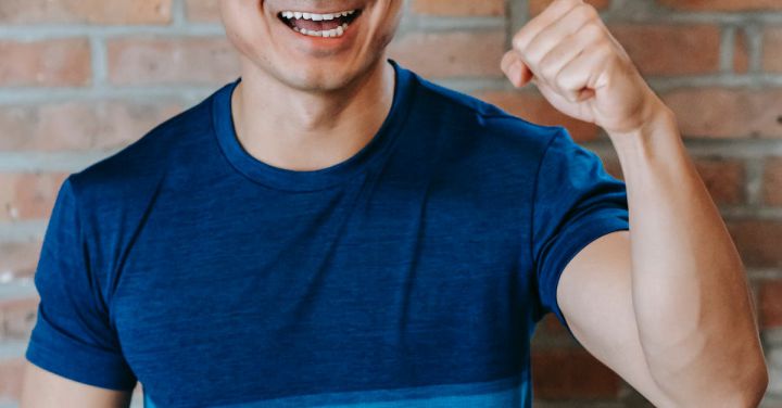 Success - Cheerful young ethnic male athlete in sportswear smiling and showing fist while standing against brick wall after successful workout in gym