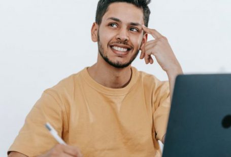 Business Strategies - Smiling young bearded Hispanic male entrepreneur thinking over new ideas for startup project and looking away dreamily while working at table with laptop and taking notes in notebook