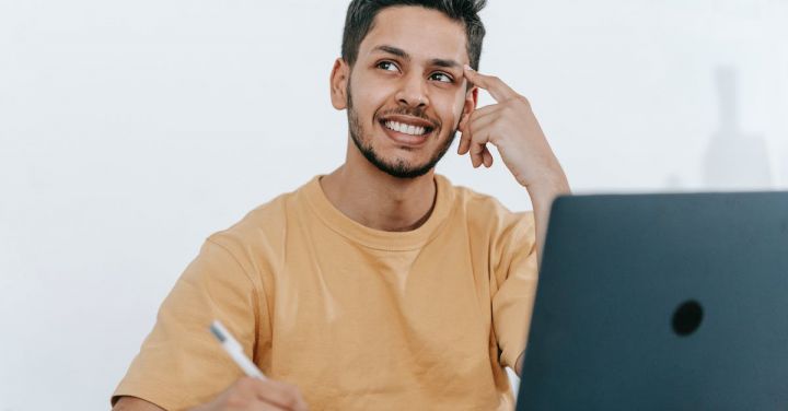 Business Strategies - Smiling young bearded Hispanic male entrepreneur thinking over new ideas for startup project and looking away dreamily while working at table with laptop and taking notes in notebook