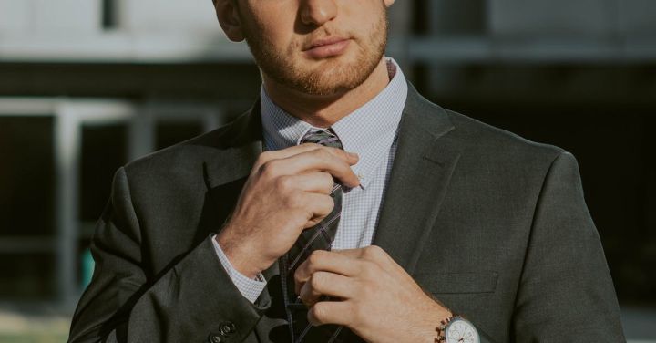 Financial Management - Serious confident male entrepreneur wearing classy suit and wristwatch holding tie while thoughtfully looking away