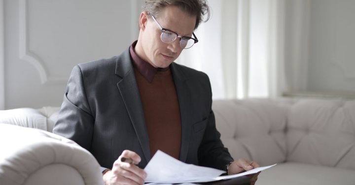 Boss - Elegant adult man in jacket and glasses looking through documents while sitting on white sofa in luxury room