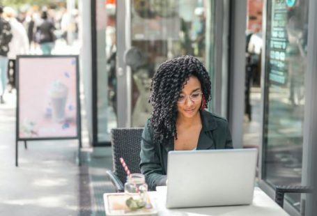 Business Connections - High angle of pensive African American female freelancer in glasses and casual clothes focusing on screen and interacting with netbook while sitting at table with glass of yummy drink on cafe terrace in sunny day