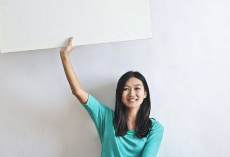 Marketing Campaigns - Cheerful Asian woman sitting cross legged on floor against white wall in empty apartment and showing white blank banner