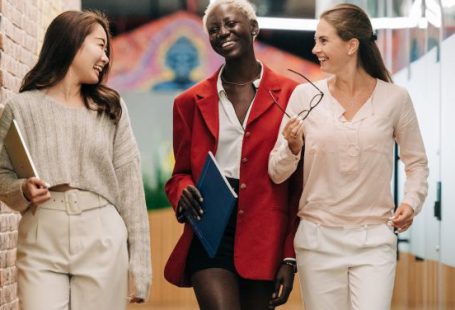 Business Strategies - Group of young glad businesswomen in trendy elegant outfits smiling and discussing business strategy in contemporary workspace