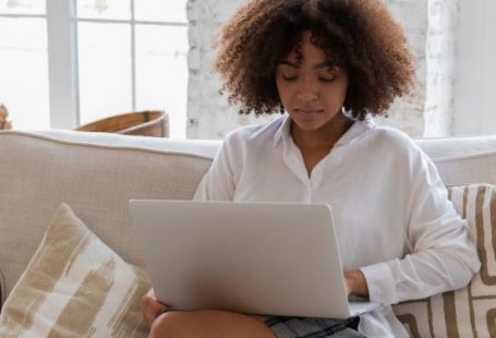 Business Connections - Full body of young stylish barefooted black female freelancer with Afro hair in casual outfit working remotely on laptop while sitting on comfortable sofa in cozy living room