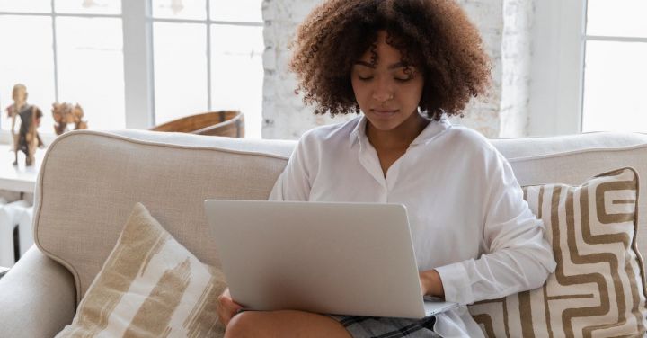 Business Connections - Full body of young stylish barefooted black female freelancer with Afro hair in casual outfit working remotely on laptop while sitting on comfortable sofa in cozy living room