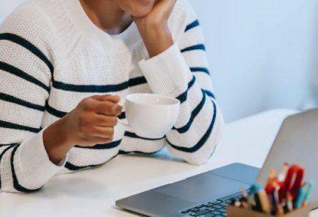Business Connections - Positive young African American female freelancer in casual clothes talking on smartphone and drinking coffee while working remotely on laptop at home