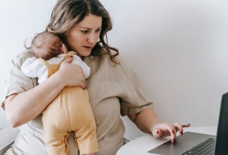Business Connections - Concentrated young female freelancer embracing newborn while sitting at table and working remotely on laptop at home