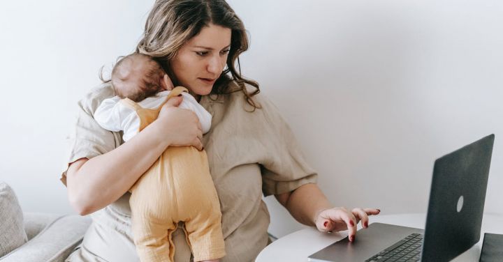 Business Connections - Concentrated young female freelancer embracing newborn while sitting at table and working remotely on laptop at home