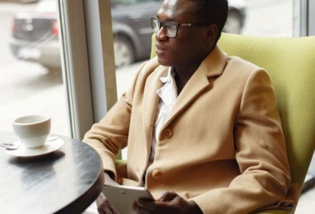 Success - Serious African American male in trendy formal suit and eyeglasses sitting on cozy chair in cafe with cup of coffee and browsing tablet
