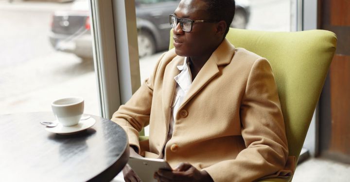 Success - Serious African American male in trendy formal suit and eyeglasses sitting on cozy chair in cafe with cup of coffee and browsing tablet